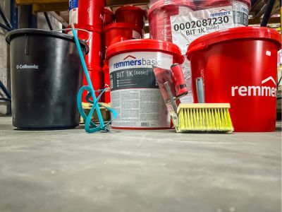 Rote und schwarze Eimer mit Werkzeugen wie Pinsel stehen auf Boden, zeigen Bauchemieprodukte im Lager.